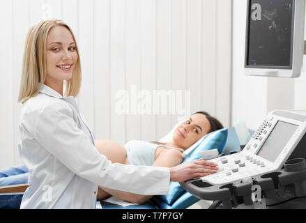 Seitenansicht der professionellen Ärztin auf Kamera und lächelnd, während in der Klinik arbeiten. Frauenarzt in weiße Uniform, Ultraschall Diagnostik in der Schwangerschaft liegende Frau auf der Couch. Stockfoto