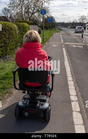 Eine ältere Dame in einem roten Mantel, in der Freiheit eines elektrischen Mobilität scooter an einer Hauptstraße, bewölkten Himmel Stockfoto