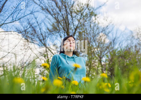 Mädchen in Hoodie sitzen auf der grünen Wiese unter Blumen und Meditieren in der Natur Hintergrund. Frau Üben der Meditation. Die Augen geschlossen. Stockfoto