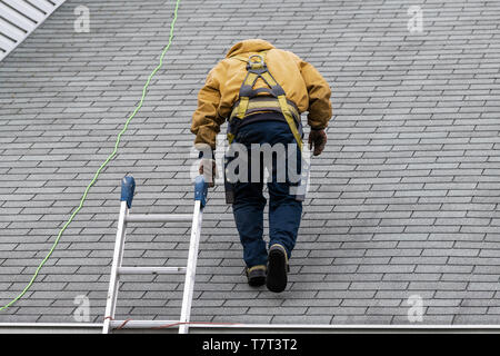 Haus während des Tages mit grauer Farbe Einfamilienhaus und Bau Mann in gelber Uniform zu Fuß auf Dachschindeln und Leiter während der Reparatur Stockfoto