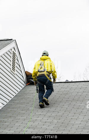 Haus während des Tages mit grauer Farbe bauen und Mann in gelber Uniform zu Fuß auf Dachschindeln und Leiter während der Reparatur Stockfoto