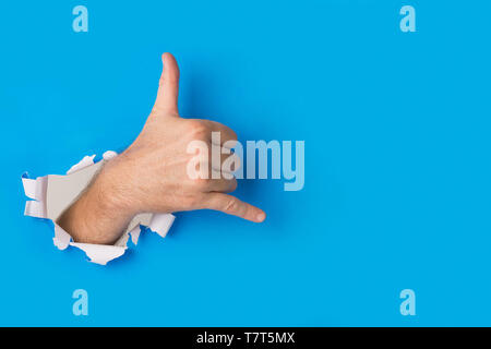 Männliche Hand reißen durch blaues Papier Hintergrund Erstellen einer Shaka Geste. Eine freundliche Hand Zeichen unter den Surfern rund um die Welt eingesetzt. Stockfoto