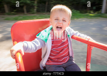 Kleinen Niedlichen kaukasischen Jungen spinnen Runde sitzen auf einem Karussell metall Stuhl in einen Spielplatz im Sommer Stockfoto