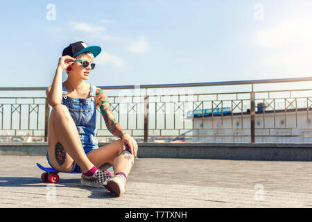 Junge alternative Mädchen skater tragen Cap und Sonnenbrille sitzen auf Penny auf der Straße zur Seite schaut nachdenklich Stockfoto