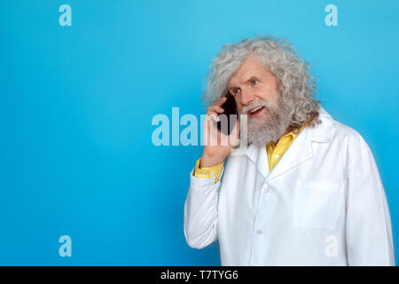 Alte langhaarigen Mann, der Doctor's Kleid studio isoliert auf blauen Wand reden auf dem Smartphone mit Freund freudigen Lächeln Stockfoto
