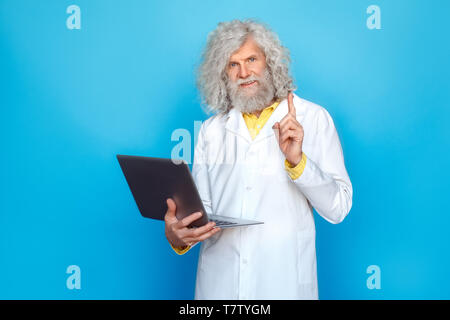 Alte langhaarigen Mann, der Doctor's Kleid Studio auf der blauen Wand holding Laptop suchen Kamera lächelt fröhlich nach oben Kreative id isoliert Stockfoto