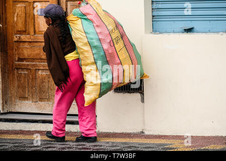 Eine alte indigene Frau, die eine große schwere Tasche auf dem Rücken bei Octaval Markt in Ecuador Stockfoto