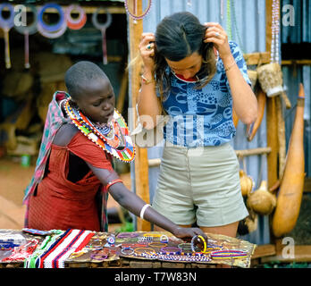 Eine weibliche Touristen versucht, auf einem nativen Perlen Halskette aus der Auswahl der Masai Kunsthandwerk an einer im Souvenir stand in Tansania, Ostafrika angezeigt. Die bunten Modeschmuck ist in der Regel von Glasperlen altmodisch wie Halsketten, Armbänder und Ohrringe. Raupe die Raupe Farben und Muster haben Bedeutung für die Maasai Frauen beaders, die in ihren Dörfern fast täglich zu tratschen sammeln und ihre einzigartigen dekorativen Schmuck zu erstellen. Stockfoto