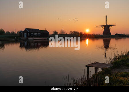 Laden Plattform am Rand mit dem ruhigen Wasser des langen Kanals während mit Blick auf eine Windmühle Reflexion in der Brennen sunrise Farbe morgen Stockfoto