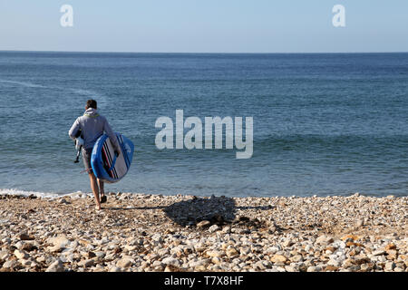 Charmouth Beach, Dorset, UK - Rückansicht eines jungen Mannes, der mit einem Paddelbrett und einem Hund unter dem Arm mit Kopierraum aufs Meer hinausgeht Stockfoto