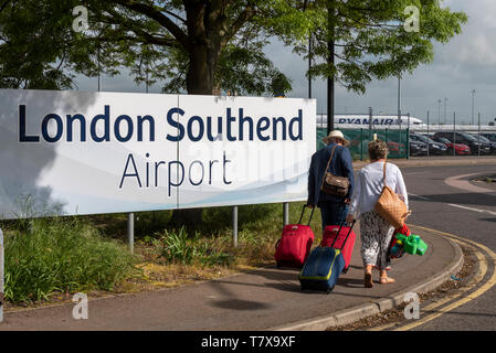 Familie von Passagieren, die am Einfahrtsschild des London Southend Airport vorbeilaufen, mit dem Flugzeug von Ryanair dahinter. Southend on Sea, Essex, Großbritannien Stockfoto