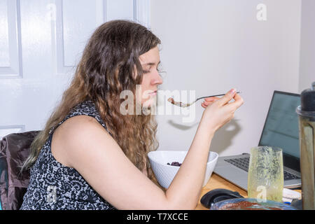 Junge Frau am Schreibtisch zu Hause sitzt mit Laptop Computer essen Haferflocken Beeren oder Eis von Blender und Smoothie cup Stockfoto