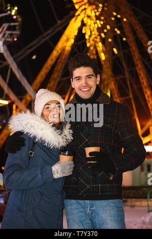 Gerne fröhliche junges Paar, das Spaß am Eislaufen Park bei Nacht, holding Tassen Kaffee zum Mitnehmen Stockfoto