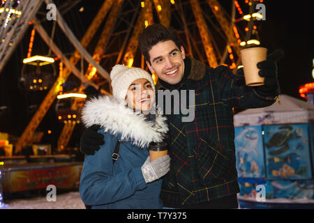 Gerne fröhliche junges Paar, das Spaß am Eislaufen Park bei Nacht, holding Tassen Kaffee zum Mitnehmen Stockfoto