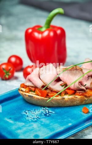 Auf einem blauen Platte des bruschet mit Rindfleisch und Tomaten, daneben ist eine rote Paprika Tomaten. Hellgrau strukturierten Hintergrund. Schöne Servieren von Speisen. Restaurant Menü Stockfoto