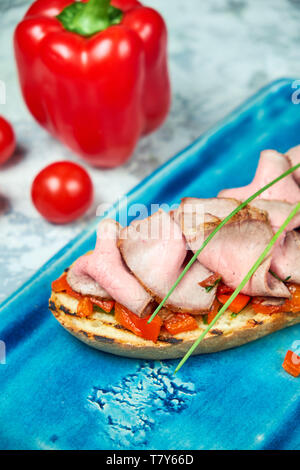 Auf einem blauen Platte des bruschet mit Rindfleisch und Tomaten, daneben ist eine rote Paprika Tomaten. Hellgrau strukturierten Hintergrund. Schöne Servieren von Speisen. Restaurant Menü Stockfoto