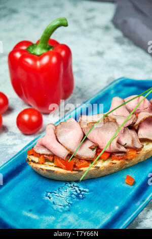 Auf einem blauen Platte des bruschet mit Rindfleisch und Tomaten, daneben ist eine rote Paprika Tomaten. Hellgrau strukturierten Hintergrund. Schöne Servieren von Speisen. Restaurant Menü Stockfoto