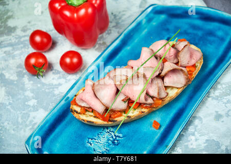 Auf einem blauen Platte des bruschet mit Rindfleisch und Tomaten, daneben ist eine rote Paprika Tomaten. Hellgrau strukturierten Hintergrund. Schöne Servieren von Speisen. Restaurant Menü Stockfoto