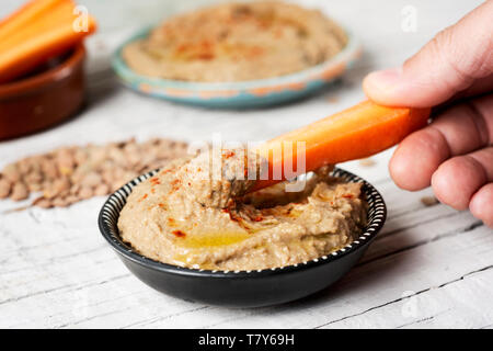 Nahaufnahme von einem Mann taucht ein Streifen von Karotte in einem hausgemachten Linse hummus gewürzt mit Paprika in einem schwarzen und weißen Teller serviert, auf einem weißen rustikalem Holz Stockfoto