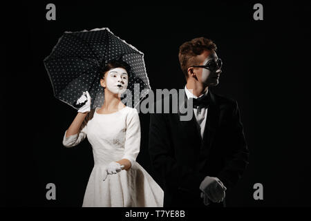 Portrait von Paar mime mit Sonnenschirm auf schwarzen Hintergrund. Mann im Smoking und Gläser und Frau im weißen Kleid. Beziehung Stockfoto