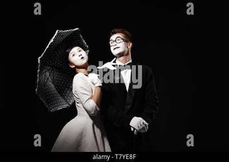 Portrait von Paar mime mit Sonnenschirm auf schwarzen Hintergrund. Mann im Smoking und Gläser und Frau im weißen Kleid. Platz für Text Stockfoto