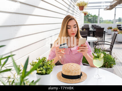 Schöne Frau in einem Restaurant Kleid, Mädchen im Sommer Cafe. Konzept des Kaufens und Abendessen Abendessen bezahlen per Kreditkarte. In der Hand des Telefons ist ein Stockfoto