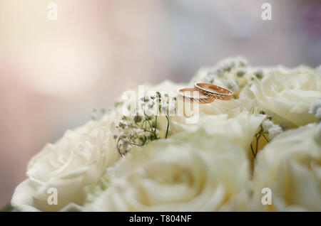 Gold Trauringe liegen auf dem weißen zarten Rosen im Sonnenlicht. Stockfoto