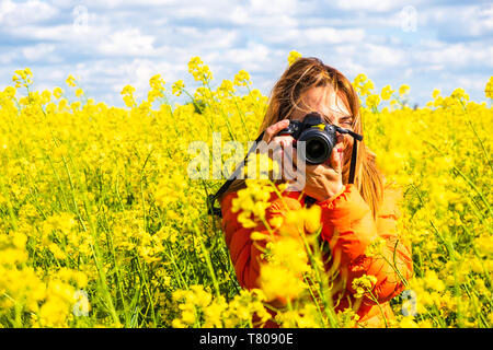 Junge Frau Fotograf mit einer DSLR, trug den orange Jacke, dauert ein Bild in einem Raps Feld, ländliche Landschaft, Osteuropa, Rumänien - Kopieren Stockfoto