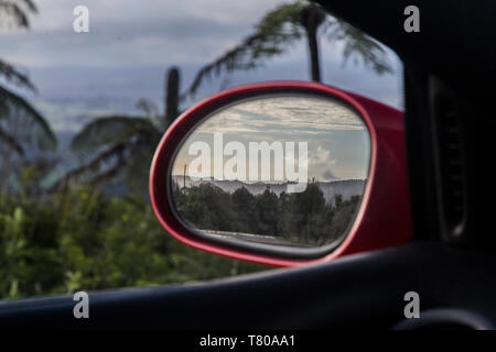 Blick in den Rückspiegel eines roten sports Racing Car. Spiegel ist im Fokus und zeigt Wolken über einem Bush Szene aus Der Waitakere Ranges in Auckland. Punga Stockfoto