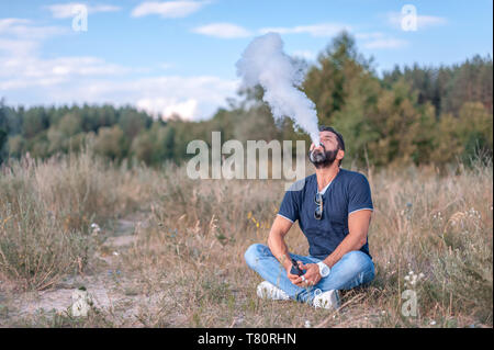 Brutale vape Mann halten und Rauchen seine elektronische Rauch Gerät blasen einen Stream von Rauch ausbreiten. Stilvolle Mann. Stockfoto