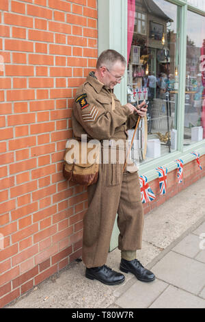 Woodhall Spa 1940 Weltkrieg zwei Festival Re-enactment Wochenende, Soldat im Zweiten Weltkrieg Schlacht gekleidet lehnte sich gegen eine Wand mit einem Smart Phone. Stockfoto