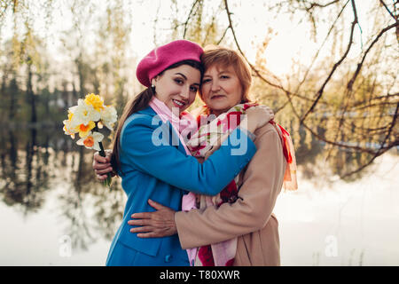 Ältere Mutter mit Blumen und ihre erwachsene Tochter umarmen von Spring River. Muttertag Konzept. Die Werte der Familie Stockfoto