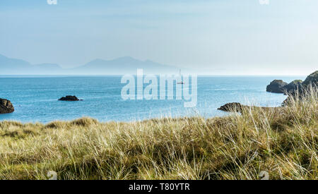 Ein Segelboot mit den Bergen der Llyn Halbinsel, die im Hintergrund von llanddwyn Island, Anglesey, North Wales, UK gesehen. Stockfoto