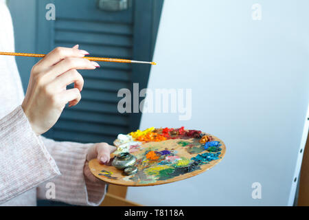 In der Nähe der weiblichen Händen in einem rosa Pullover mit einem Besen und eine Palette mit bunten Acrylfarben gegen eine weiße Leinwand, selektiven Fokus Stockfoto