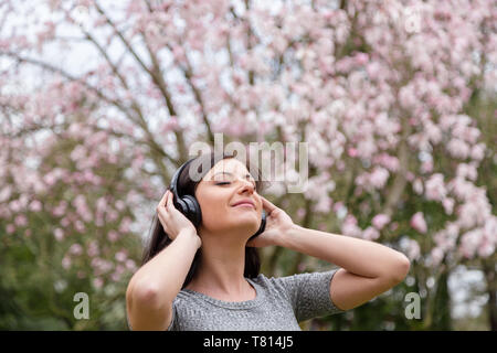 Junge Frau hören die Musik auf kabellosen Kopfhörern in einem Park mit Kirschblüten Bäume. Stockfoto