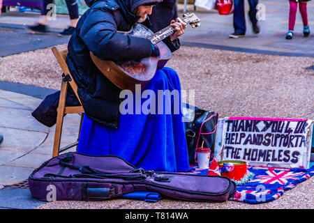 Eine obdachlose Gaukler mit einem Schild mit einer Gitarre in einer belebten Einkaufsstraße in Oxford, Großbritannien spielt, im Winter. Dezember 2018. Stockfoto