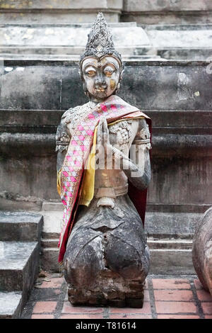 Alte vintage scaried Stein Buddhismus Statue; Leuchten gelbe Auge, hebt seine Hand und knie dich auf die Bühne, schauen auf den Boden. Chiangrai, Thailand. Stockfoto