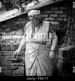 Schwarz-weiße vorne, Frau mittleren Alters, gekleidet in Kostüm als alte WW2-Hausfrau in Kopftuch & Overalls von Brick Outhouse, UK-Veranstaltung der vierziger Jahre. Stockfoto