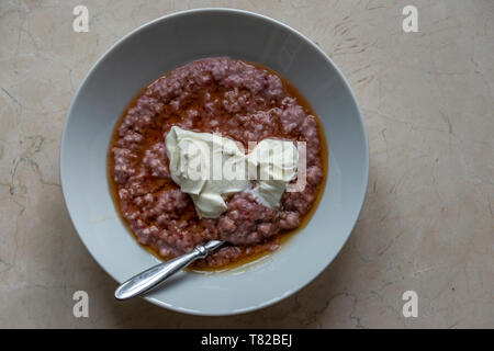 Eine Vogelperspektive auf einem Teller Porridge mit silbernen Löffel, mit einem natürlichen Joghurt nachfüllen. Stockfoto