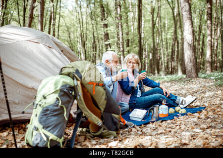 Senior Paar sitzen zusammen auf dem Campingplatz mit Zelt und Rucksack, die Natur genießen und trinken Kaffee in den Wald Stockfoto