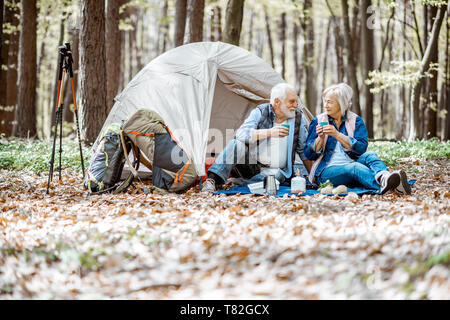 Senior Paar sitzen zusammen auf dem Campingplatz mit Zelt und Rucksack, die Natur genießen und trinken Kaffee in den Wald Stockfoto