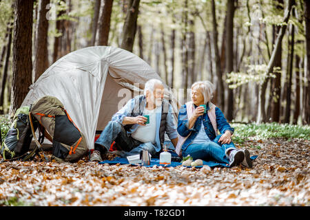 Senior Paar sitzen zusammen auf dem Campingplatz mit Zelt und Rucksack, die Natur genießen und trinken Kaffee in den Wald Stockfoto