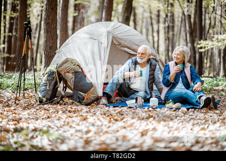 Senior Paar sitzen zusammen auf dem Campingplatz mit Zelt und Rucksack, die Natur genießen und trinken Kaffee in den Wald Stockfoto