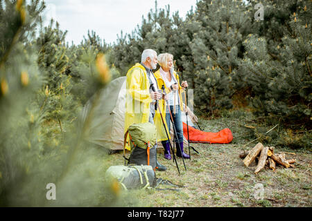 Senior Paar in gelben Regenjacken auf dem Campingplatz mit Zelt und Kamin in der junge kiefernwald Stockfoto