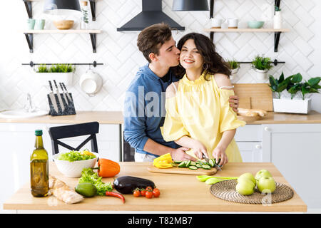 Junge nette lächelnde Paar kochen gemeinsam in der Küche zu Hause. Eine junge Frau schneiden frisches Gemüse auf einem Holz- Salat. Stockfoto