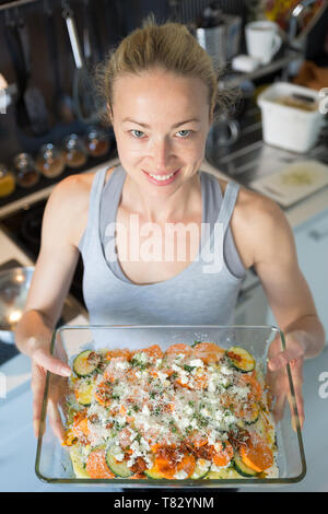 Lächelnde junge gesunde Frau halten und stolz zeigen Glas backen Versuchen mit Reihe vegetarischer Teller Zutaten, bevor Sie es in den Ofen. Stockfoto