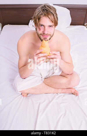 Frische Backwaren Produkt. Man bärtige hübscher Kerl essen Frühstück im Bett. Französisches Frühstück stereotyp. Kerl hält croissant Bett im Schlafzimmer oder im Hotelzimmer zu sitzen. Der Mensch isst Croissant er Backwaren mag. Stockfoto