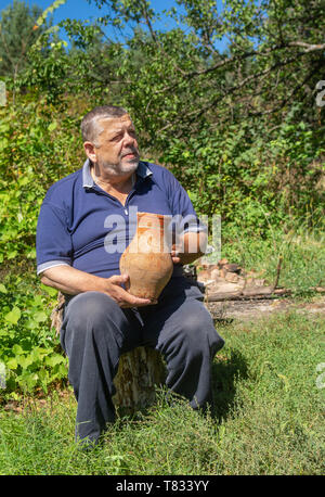 Outdoor Portrait von Ukrainischen senior Bauer sitzt auf einem Anmelden und unter einem alten Ton Krug Stockfoto