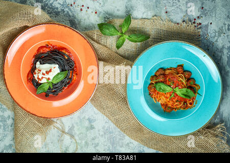 Blau Teller mit Nudeln und Fleisch Kugeln, orange Platte mit Tinte vom Tintenfisch Tinte. Graues Licht strukturierten Hintergrund. Schöne Servieren von Speisen. Restaurant Menü Stockfoto