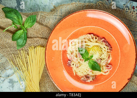 Pasta Carbonara mit Ei und Basilikum auf einem orangefarbenen Platte. Grau strukturierten Hintergrund mit beiger Stoff. Schöne Servieren von Speisen. Restaurant Menü Stockfoto